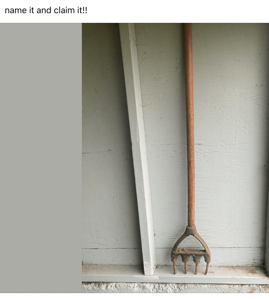 Rusty garden rake with a wooden handle leaning against a light gray wooden wall and a white beam.