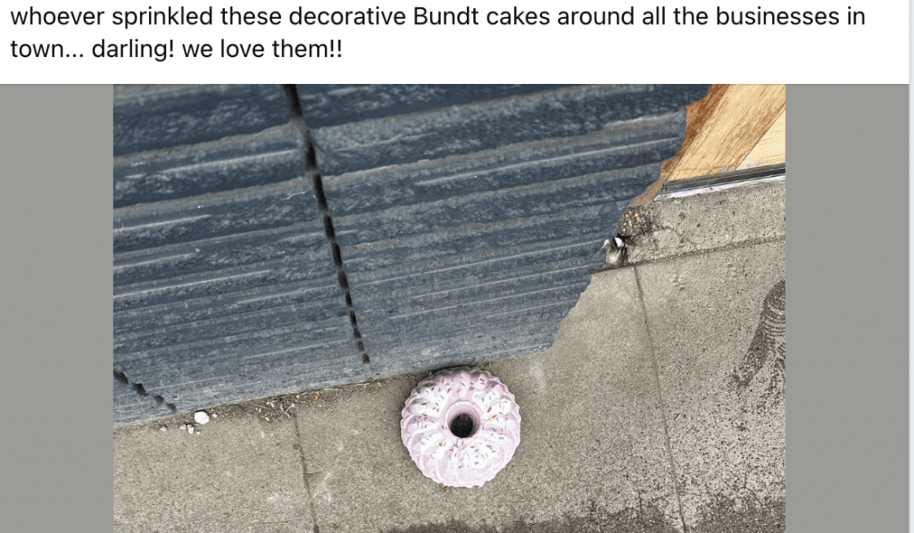 Pink frosted Bundt cake resting on a concrete sidewalk next to a gray building wall with wood trim in the background.