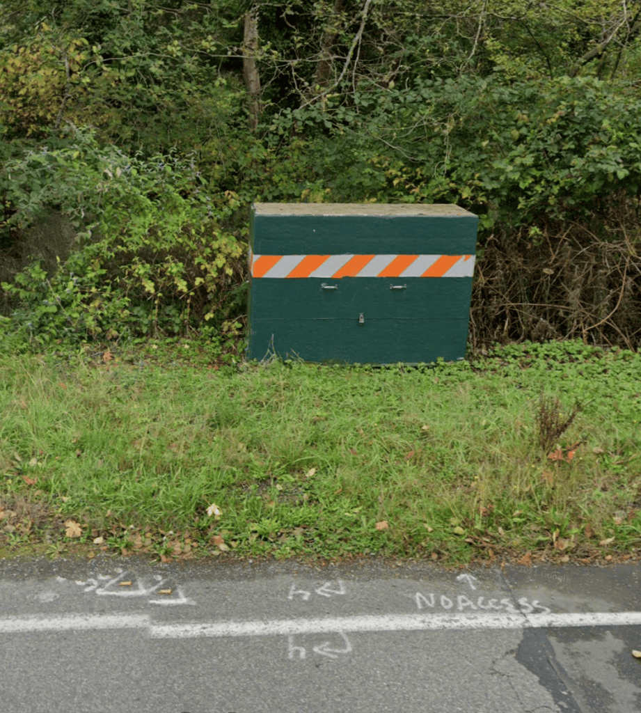 Green utility box with orange-and-white diagonal hazard stripes beside a grassy roadside, with dense shrubs in the background.
