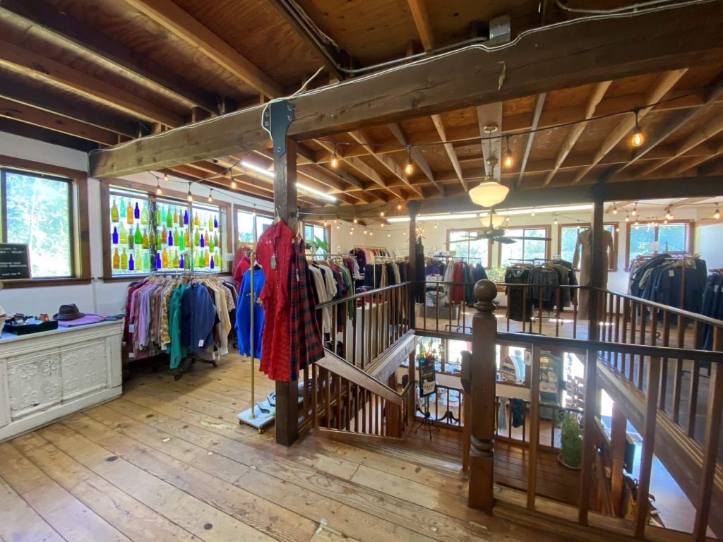 Agnes and Chloe | Vashon Maury Island Rustic clothing store interior with exposed wooden beams, hanging lights, and rows of jackets and shirts on racks near a balcony railing.