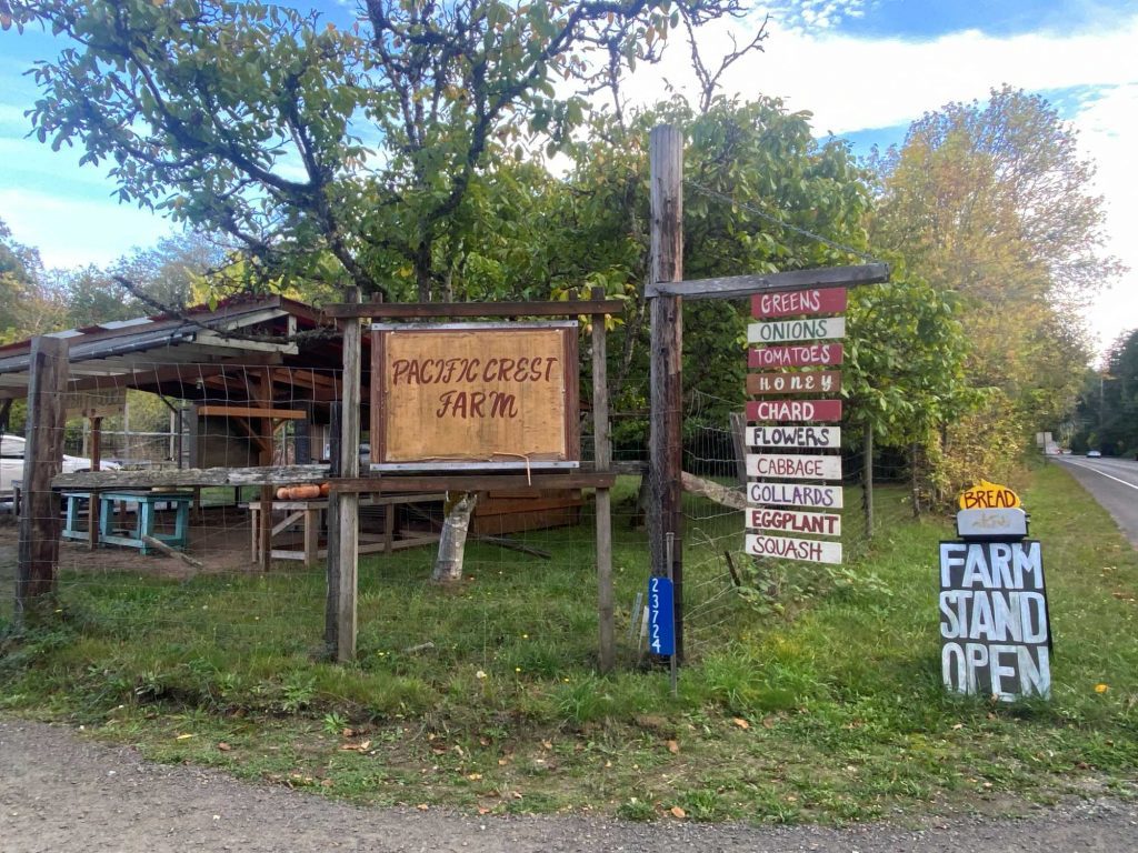 Farm entrance with a wooden sign reading 'Pacific Crest Farm' and a vertical list of produce: greens, onions, tomatoes, honey, cabbage, squash, etc., near a road.